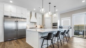 A modern kitchen with white cabinetry, stainless steel appliances, and a bar area with stools. - New luxurious home - hot tub, firepit, & trails - Walk or ride to downtown! (Bentonville)