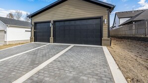 A two-car garage attached to a building with a brick driveway and a wooden fence. - New luxurious home - hot tub, firepit, & trails - Walk or ride to downtown! (Bentonville)