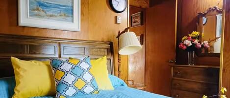 A bedroom with a wooden headboard, a bed with blue bedding and yellow pillows, a wall clock, a framed painting, and a mirror with flowers.