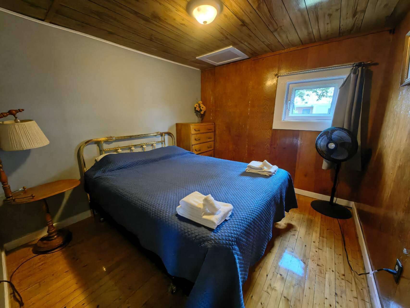 A bedroom with a bed, a nightstand, a lamp, a dresser, a fan, and a window.