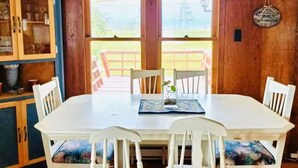 A dining area with a white table and chairs, a lace window valance, and a view of a wooden deck outside. - Pine Cottages (Stanhope)