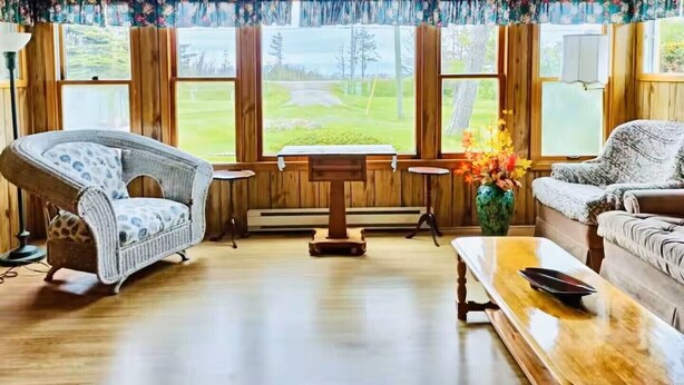 A cozy room with wooden walls and floors, a wicker chair, a sofa, a small table, and a view of a green landscape through large windows. - Pine Cottages (Stanhope)