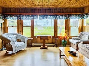 A cozy room with wooden walls and floors, a wicker chair, a sofa, a small table, and a view of a green landscape through large windows. - Pine Cottages (Stanhope)