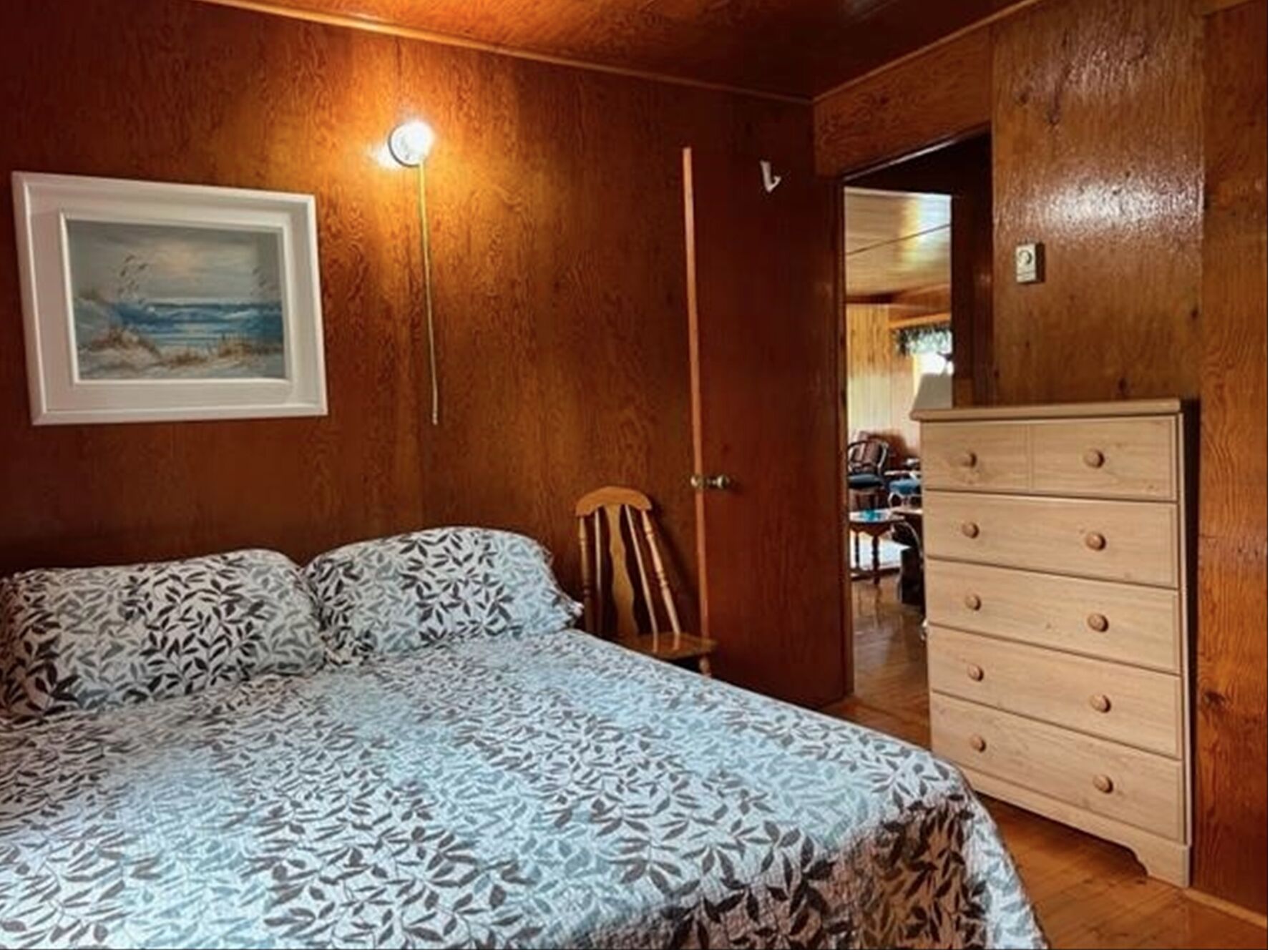Un dormitorio con una colcha estampada, un aparador, una silla y un cuadro en la pared.