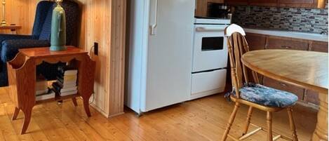 A compact kitchen with wooden walls, a white refrigerator, a microwave, a stove, and a dining table with a chair.