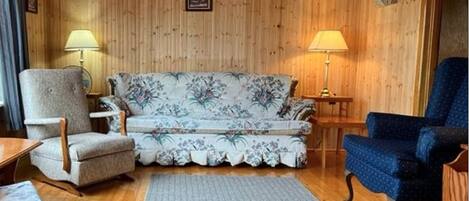 Een gezellige kamer met houten wanden, een bloemenbank, twee fauteuils, een kleed en twee lampen.