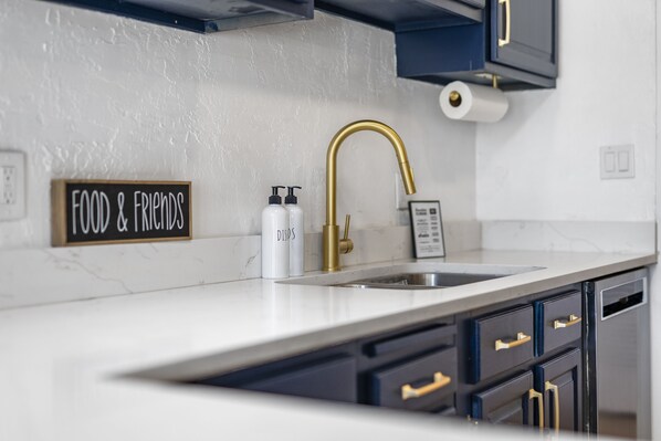A kitchen with a white countertop, a gold faucet, and dark cabinets with gold handles. There is a sign that reads "FOOD & FRIENDS" and two bottles labeled "DISOS".