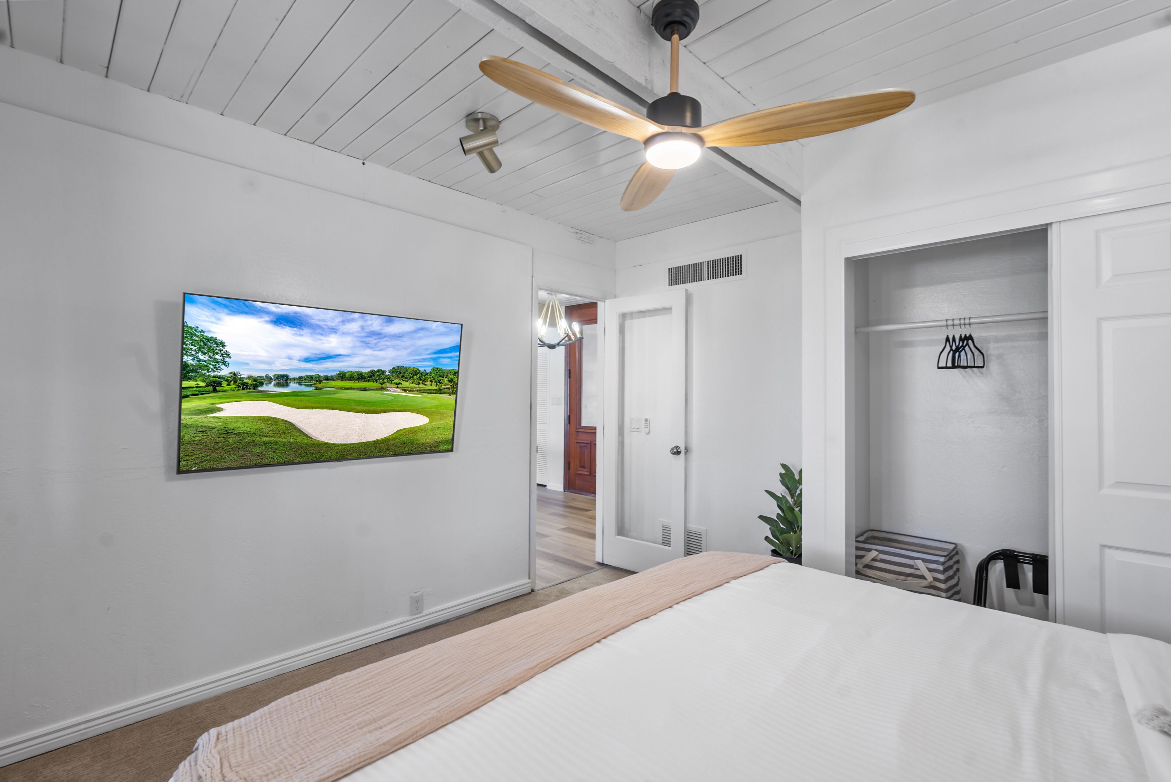 Un dormitorio con una cama grande, un ventilador de techo, un televisor con pantalla mostrando un campo de golf y una puerta que da a otra habitación.