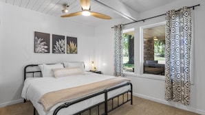 Une chambre à coucher comprenant un lit, un ventilateur de plafond, une fenêtre avec des rideaux à motifs et des œuvres d’art accrochées au mur.