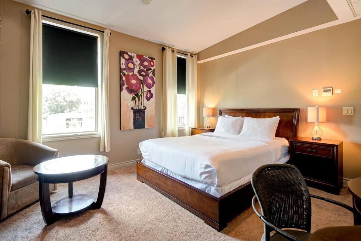 A hotel room with a large bed, a chair, a small table, a painting, and a window with curtains.