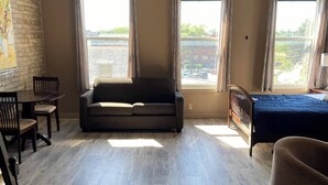 A spacious living area with a sofa, a bed, and a dining table. There are large windows with curtains, a brick wall with a painting, and a wooden floor. - The Avonlea Inn (Stratford)