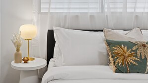 A neatly made bed with white bedding, a decorative pillow with a palm tree design, and a bedside table with a lamp and a vase of dried flowers. - Cute & Compact, pet friendly apartment easy access to Alex or Mooloolaba beaches (Alexandra Headland)