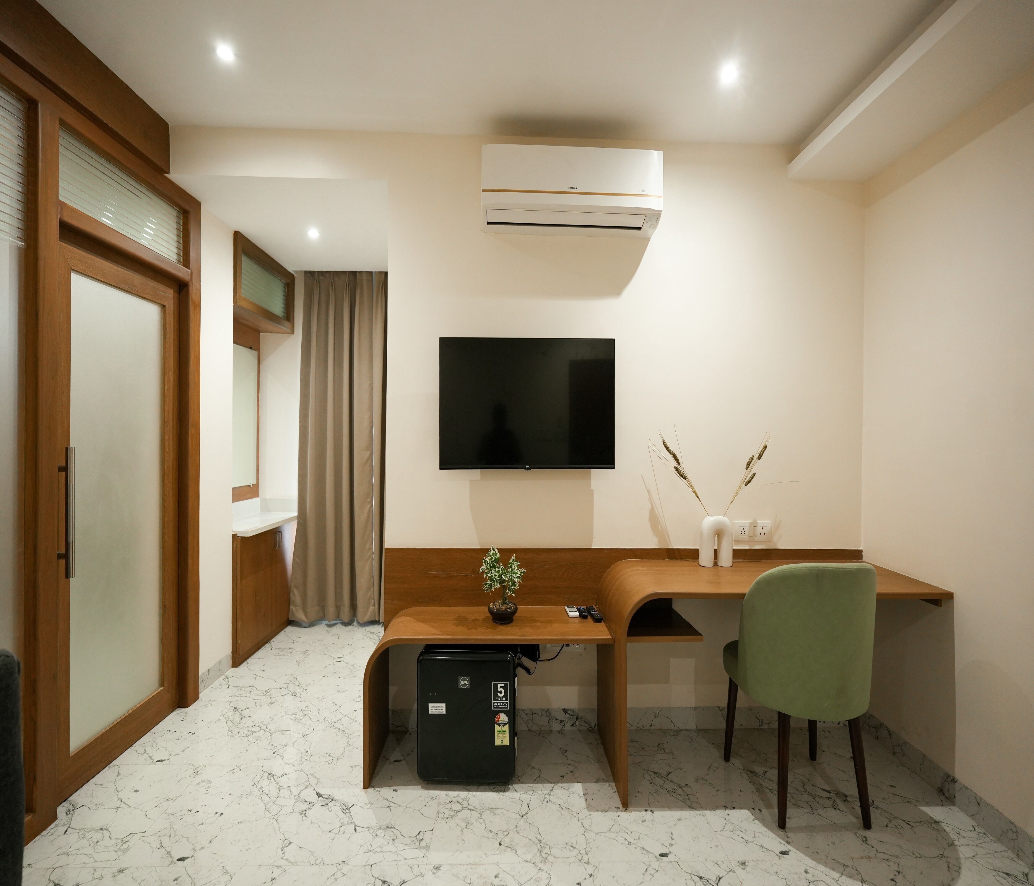 A modern room with a wooden desk, a green chair, a flat-screen TV, an air conditioner, and a small refrigerator.