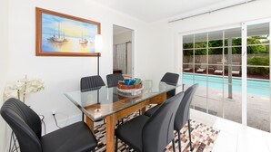 A modern dining area with a glass table and black chairs, a painting of boats on the wall, and a view of a swimming pool outside. - Sunshine State of Mind House (Jensen Beach)