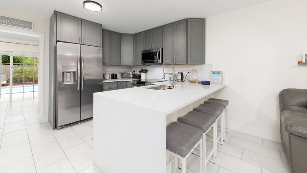 A modern kitchen with grey cabinets, a stainless steel refrigerator, a microwave, and a white island with bar stools. - Sunshine State of Mind House (Jensen Beach)
