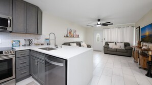 A modern kitchen with grey cabinets, a white countertop, and stainless steel appliances. There is a dining area with a grey sofa and a table. - Sunshine State of Mind House (Jensen Beach)