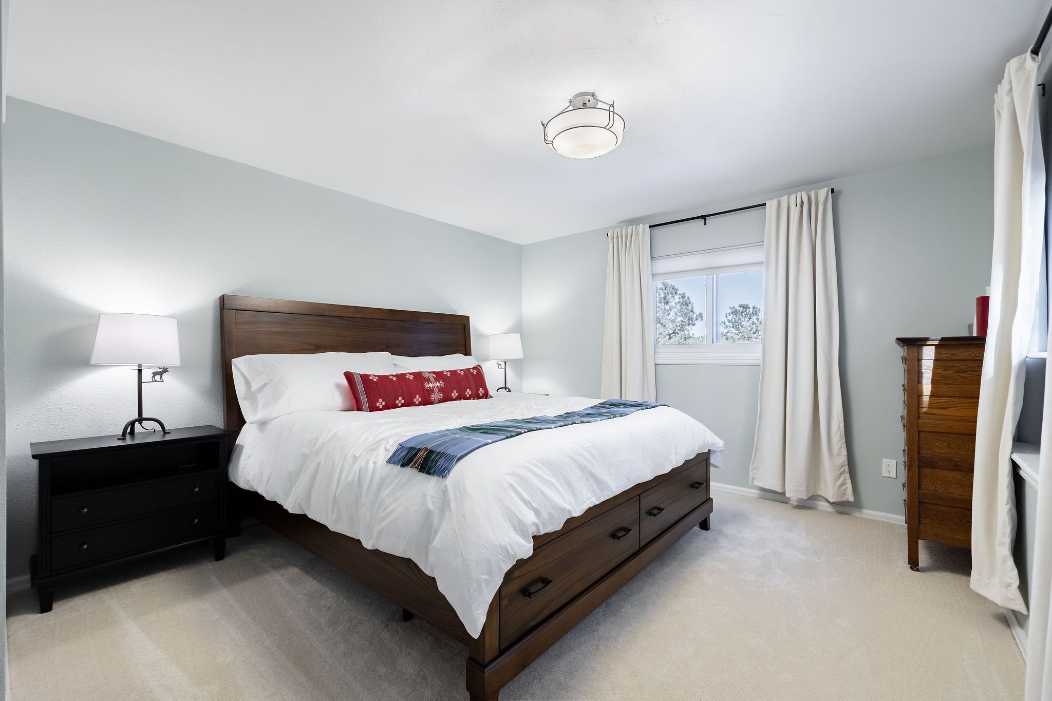Une chambre à coucher avec un lit en bois, du linge de lit blanc, une table de chevet avec une lampe, une commode et une fenêtre avec des rideaux.