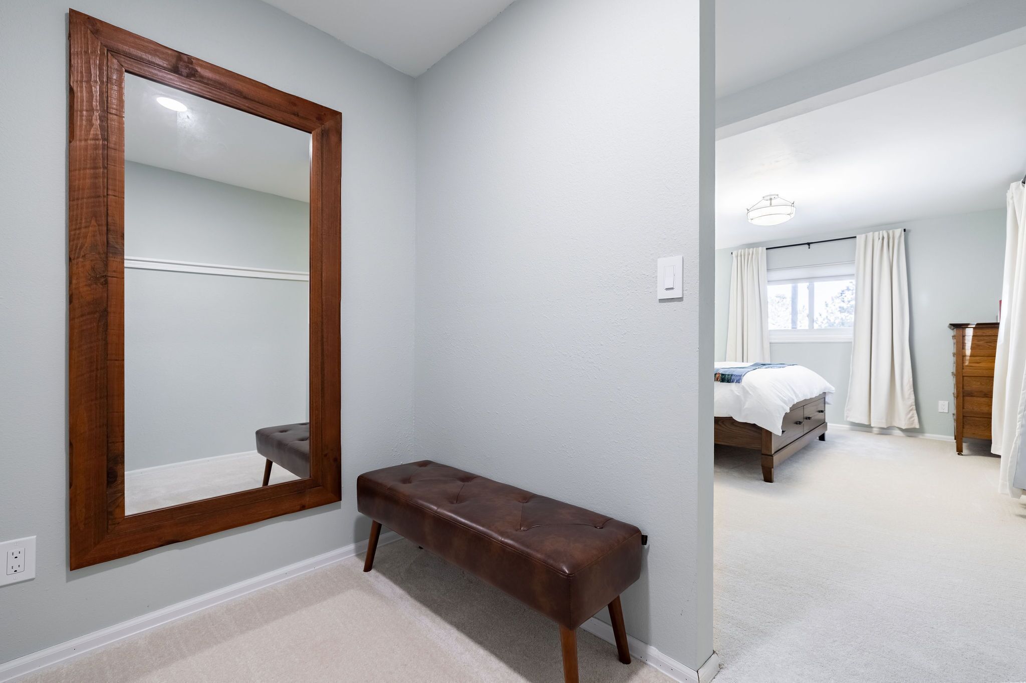 Un dormitorio con un espejo grande, una banca, una cama y una ventana con cortinas.