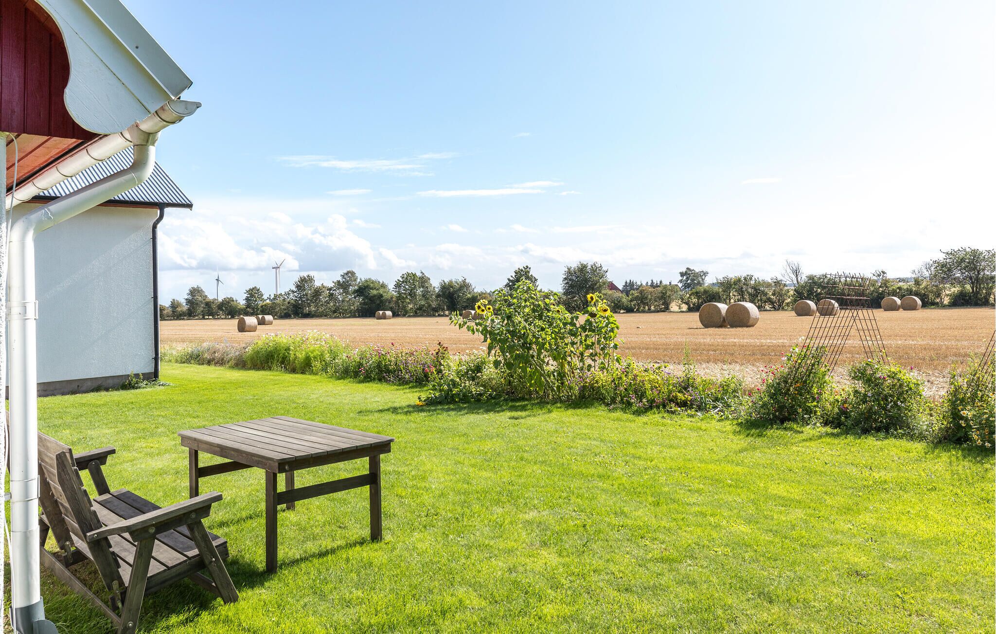 Un espace extérieur verdoyant avec une table et des chaises en bois, un bâtiment blanc avec un toit rouge, et des bottes de foin en arrière-plan.