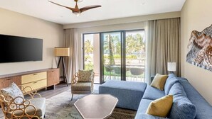 Ein modernes Wohnzimmer mit großem Flachbildfernseher, Deckenventilator, Sofa, Couchtisch und Blick auf Palmen durch große Fenster.