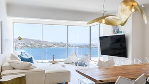 Una sala moderna con un gran ventanal con vistas a un cuerpo de agua, una mesa de comedor de madera y un televisor de pantalla plana montado en la pared.