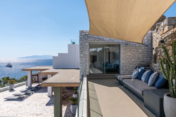 A modern outdoor seating area with a stone wall, a glass door leading inside, and a view of the sea.