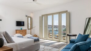A bedroom with a bed, a TV, a dresser, a sofa, and a view of a city through large windows. - Channel I in Mykonos (Mykonos)