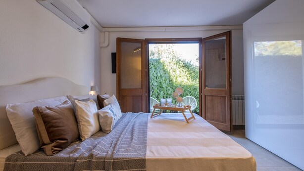 Quarto com cama, almofadas, mesa de cabeceira e uma cadeira. Há uma janela e uma porta que dá para uma área externa.