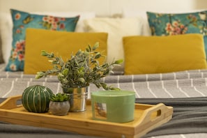 Un plateau en bois sur lequel se trouvent un pot en céramique verte, une petite plante en pot et une bougie verte, le tout posé sur un lit agrémenté d'oreillers à motifs floraux et d'une couverture à rayures.