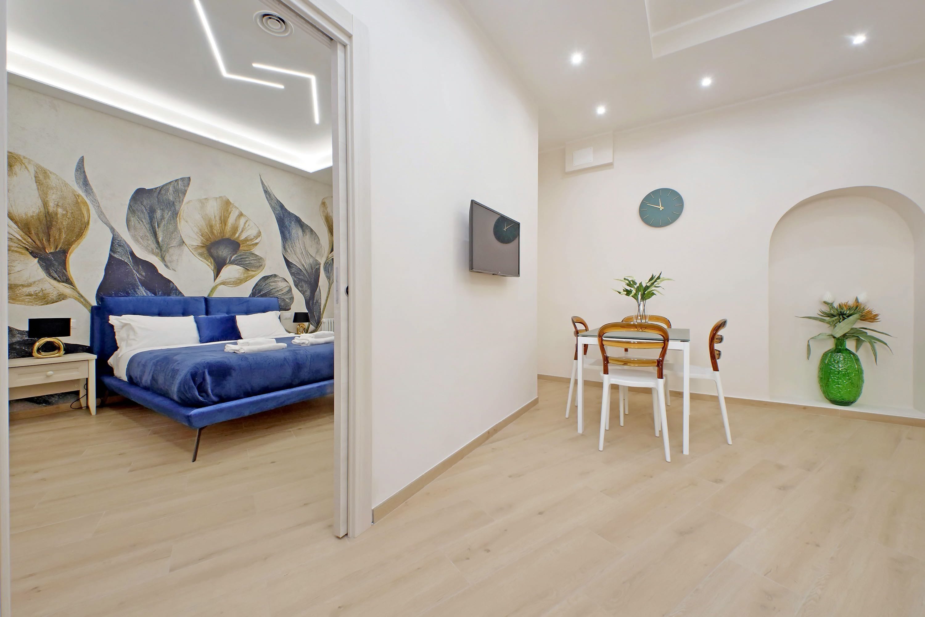 Un dormitori modern amb un llit blau, una zona de menjador amb taula i cadires, i un rellotge de paret.