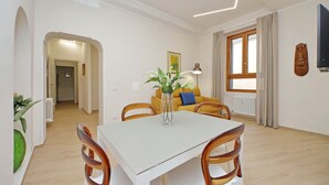 Un comedor con una mesa blanca y sillas de madera, un jarrón con flores y un sofá con un cojín azul.