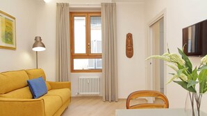 Una sala de estar con un sofá amarillo, una mesa, una silla, una lámpara, una ventana con cortinas, un radiador y un televisor.