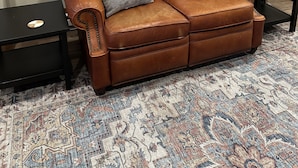 Un canapé en cuir marron agrémenté de trois coussins, un tapis à motifs et une photographie encadrée accrochée au mur.