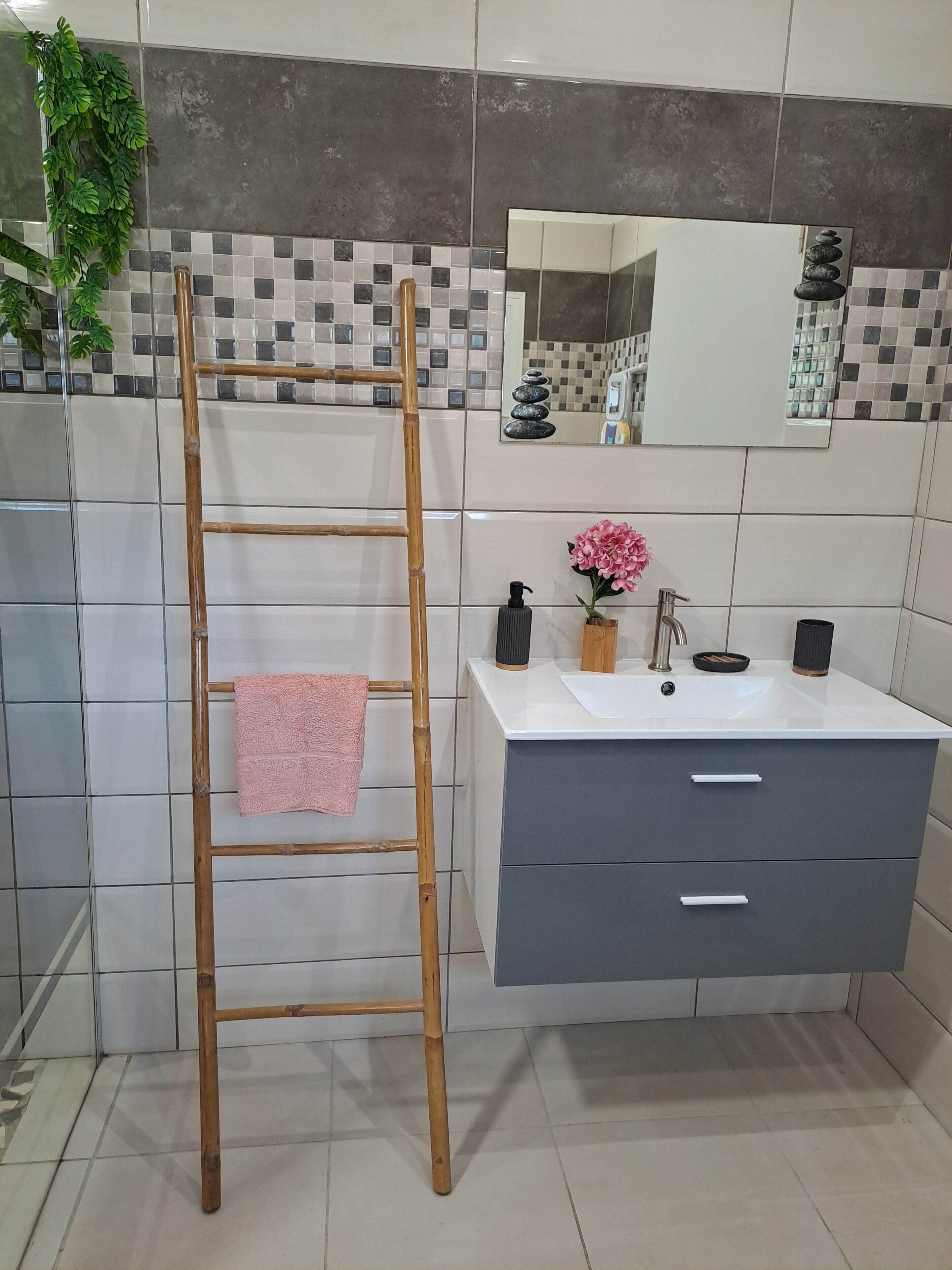 Une salle de bain avec un escabeau en bambou, une serviette rose, un lavabo avec un miroir et une pile de pierres décoratives.
