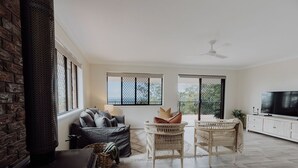A spacious living room with a fireplace, a sofa, wicker chairs, a TV, and large windows. - Indooor & Outdoor Fireplace | Acreage | Ocean View | 10min to Australia Zoo (Mount Mellum)