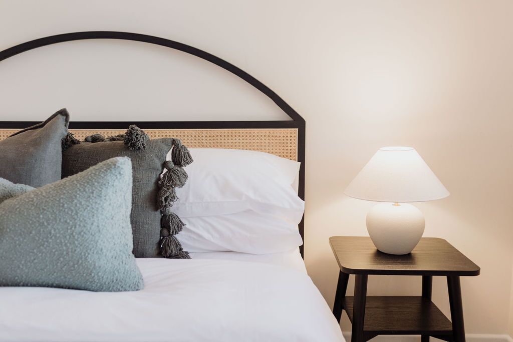 A neatly made bed with white linens and decorative pillows, a bedside table with a lamp, and a simple headboard.