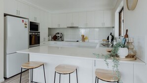 A modern kitchen with white cabinetry, a central island with stools, a refrigerator, and a coffee machine. - Indooor & Outdoor Fireplace | Acreage | Ocean View | 10min to Australia Zoo (Mount Mellum)