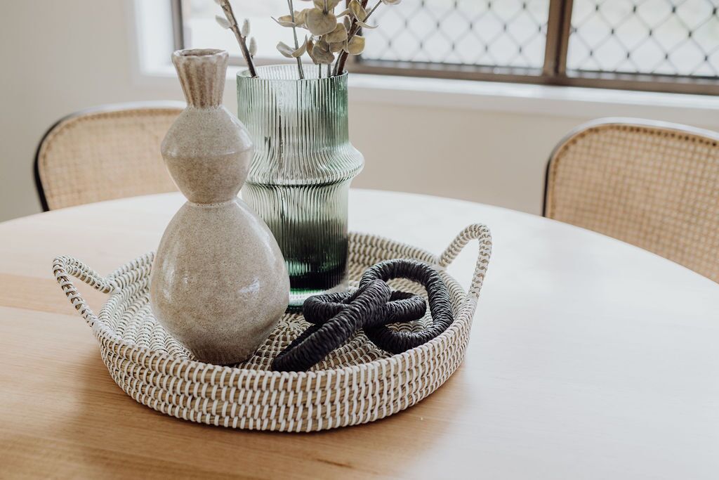 A round table with a woven tray holding a stone vase, a metal vase with branches, and a black cord.