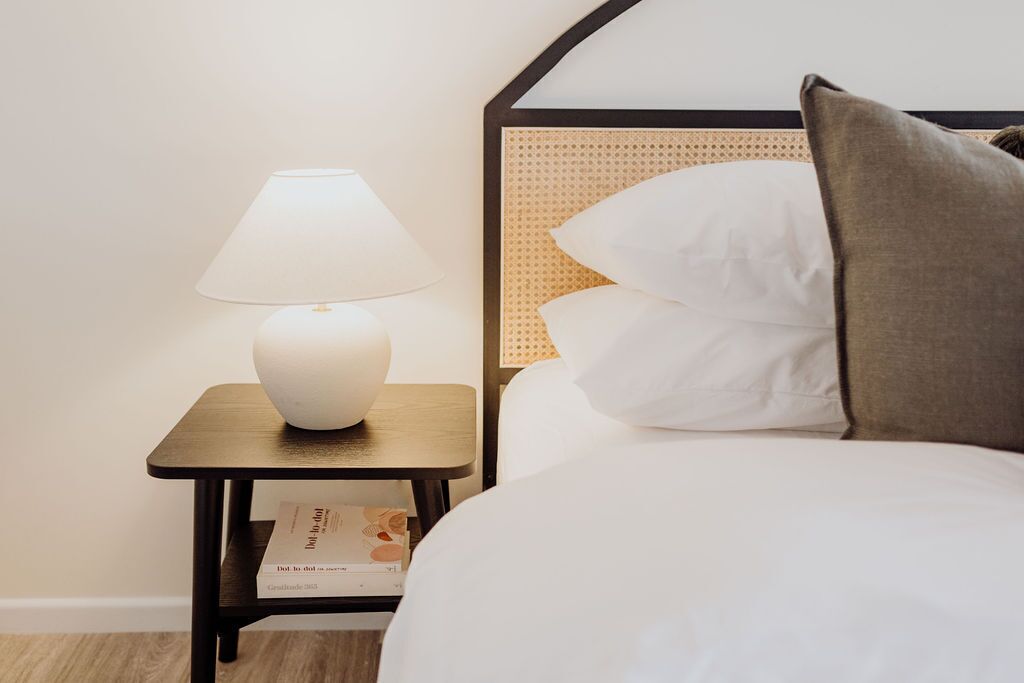 A neatly made bed with white bedding and a single pillow, a bedside table with a lamp, and a book on the table.