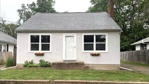 A small, single-story house with a white exterior and a grey roof. It has a front door, two windows on the front facade, and a small front yard with a brick border. - Pekin Cottage (Pekin)