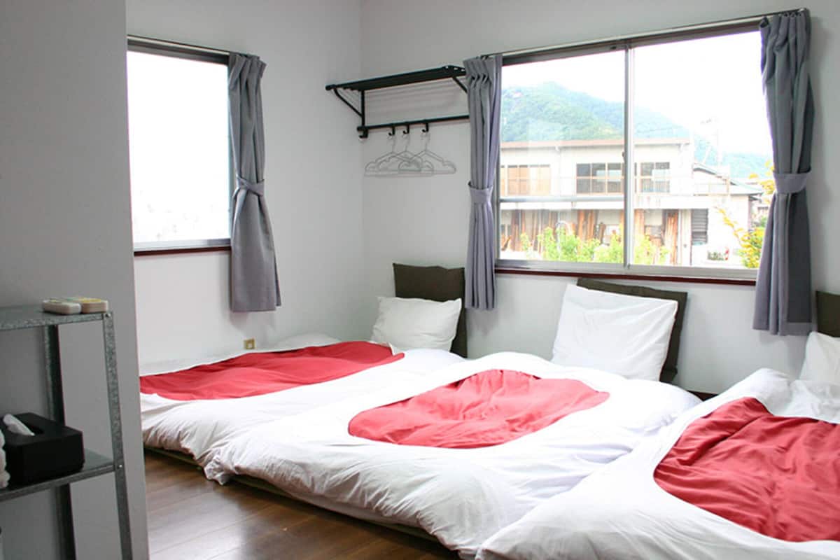 A room with two beds, a window with a view of buildings and greenery, a shelf with items, and a wall-mounted rack with hangers.