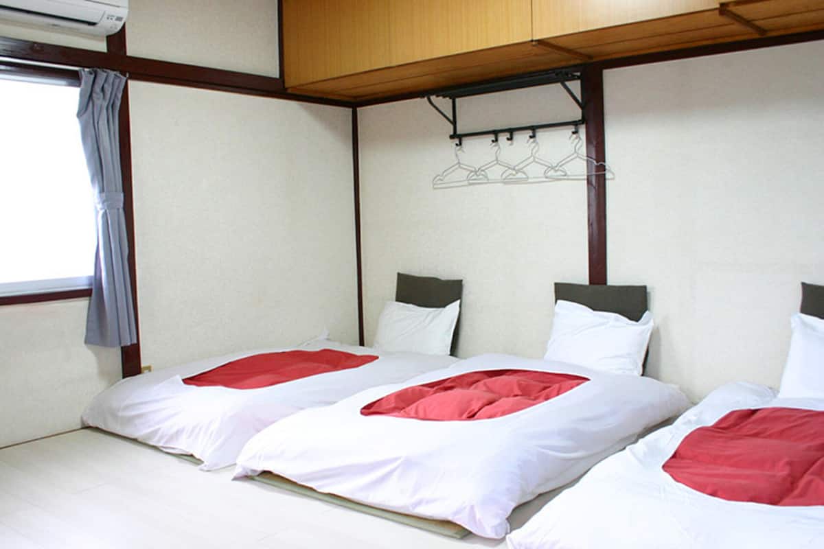 A room with two beds, each with white bedding and red accents, a window with a curtain, and a wall-mounted coat rack.
