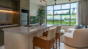 A modern kitchen with a central island, bar stools, and a large window offering a view of greenery outside. - Vesna Townhouse 1 (Canggu)