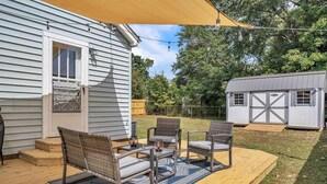 Une terrasse avec des meubles en osier et un tapis bleu, une maison blanche avec un garage et une terrasse en bois.