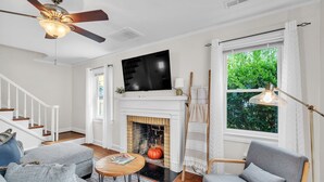 Un salon avec une cheminée, un téléviseur, un ventilateur de plafond et une grande fenêtre avec des rideaux.