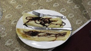 En tallerken med to pandekager dryppet med chokoladesauce, en gaffel og en kniv på et bord med et mønstret borddække.