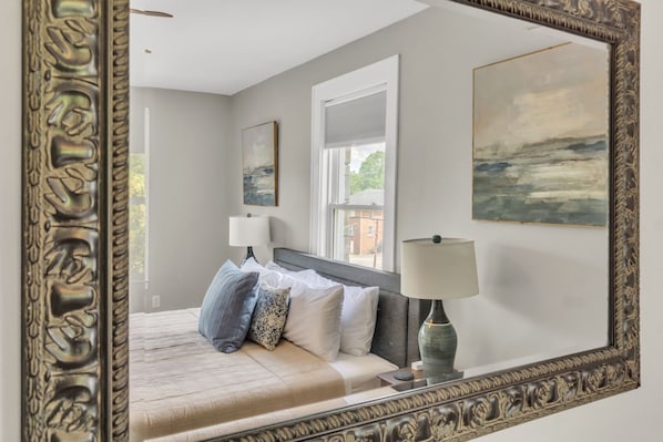 A bedroom with a large bed, two bedside lamps, a window with blinds, and a painting on the wall.