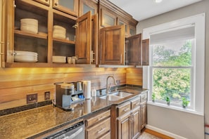 A kitchen with wooden cabinets, a granite countertop, a sink, and a window with a view of trees.