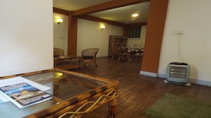 A room with a glass-top table, wicker chairs, a dining table with chairs, a standing fan, and a green rug.