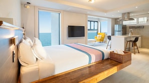 Una habitación de hotel moderna con una cama grande, televisión, balcón con vista al mar, una pequeña cocina y un área de descanso.